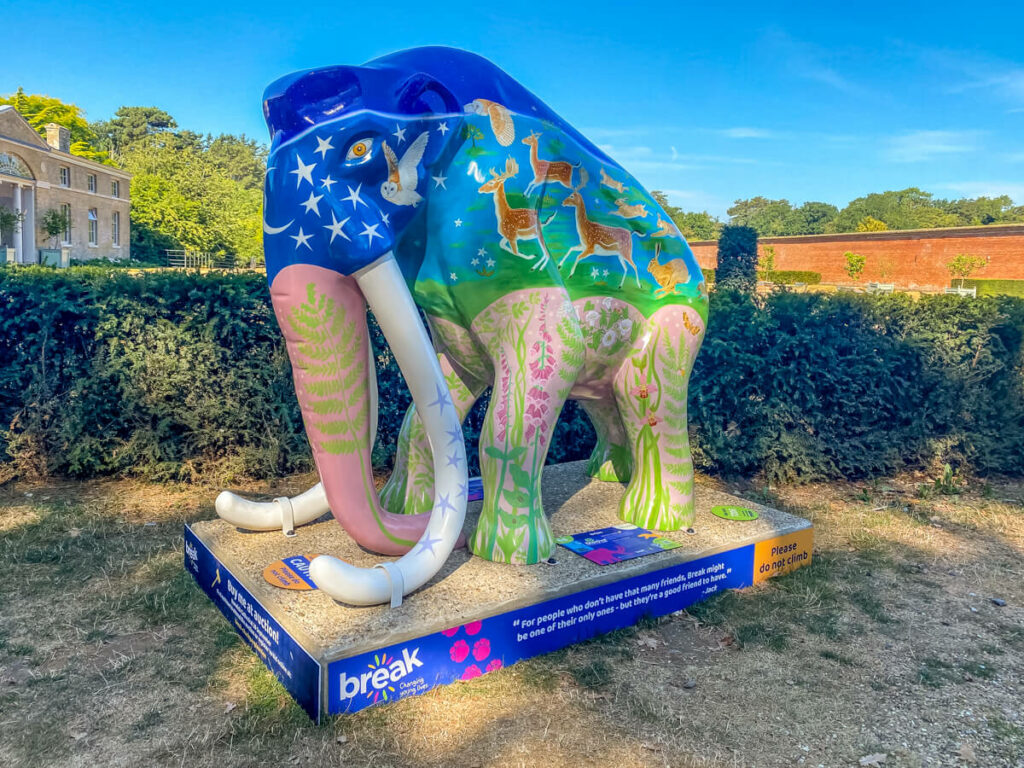 colourful mammoth with stars on its head and pink trunk and legs in front of a hedge