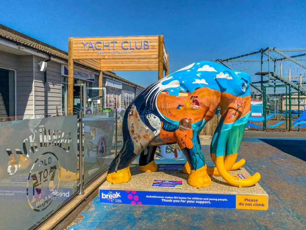 Mammoth with blue skies and clouds painted on the top, then sandy scene in the middle and seal by the tail in front of sign that says Yacht Club