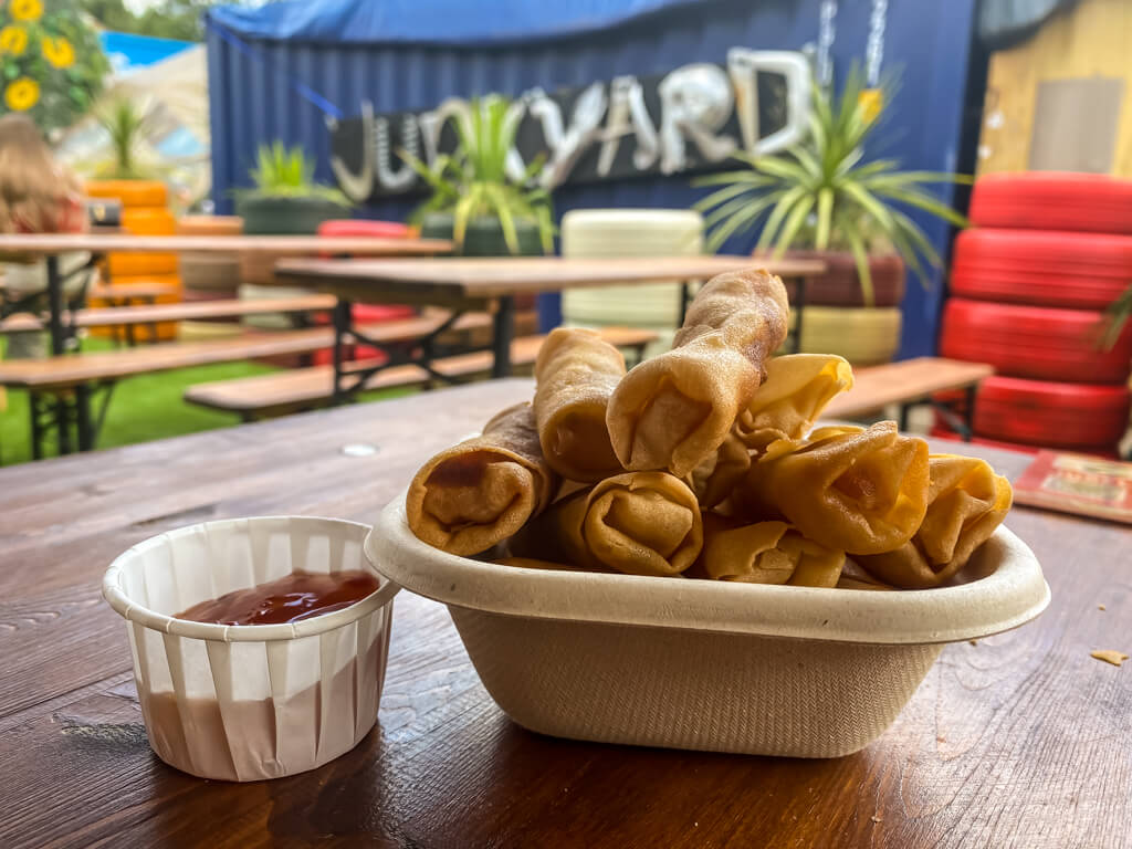 small cup of sauce, bowl of spring rolls in front of tables and you can see the word "junkyard" on the wall