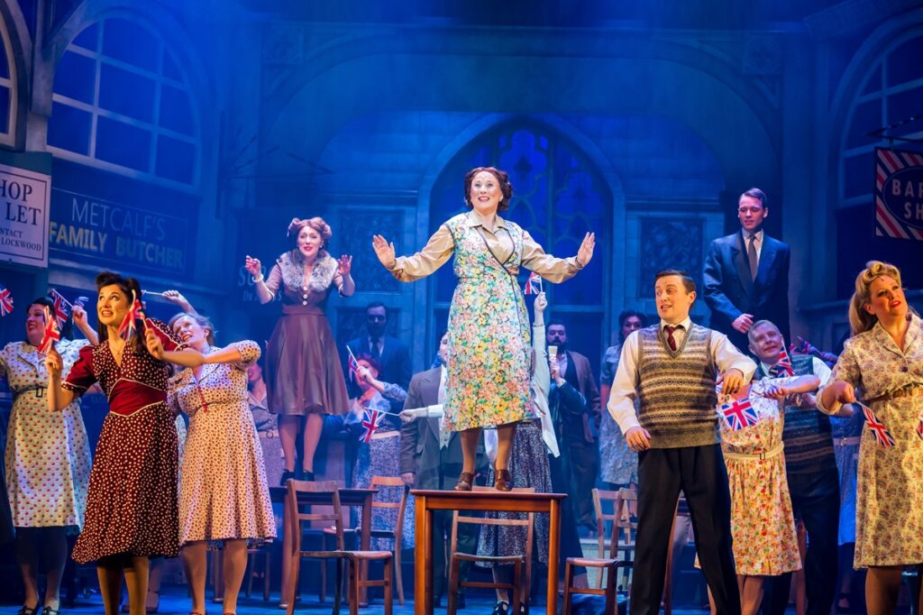 Joyce stands on a table to sing with the rest of the cast singing and dancing around her